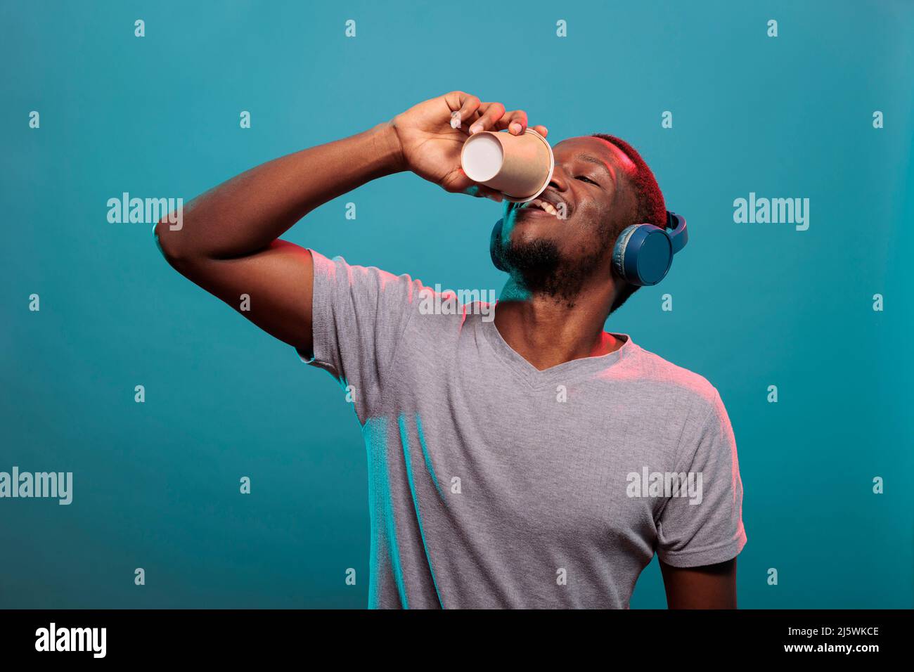 Excited man drinking coffee from cup and wearing headphones to listen to  radio music on audio gadget. Casual person with wireless earphones enjoying  drink and mp3 sound to relax Stock Photo -