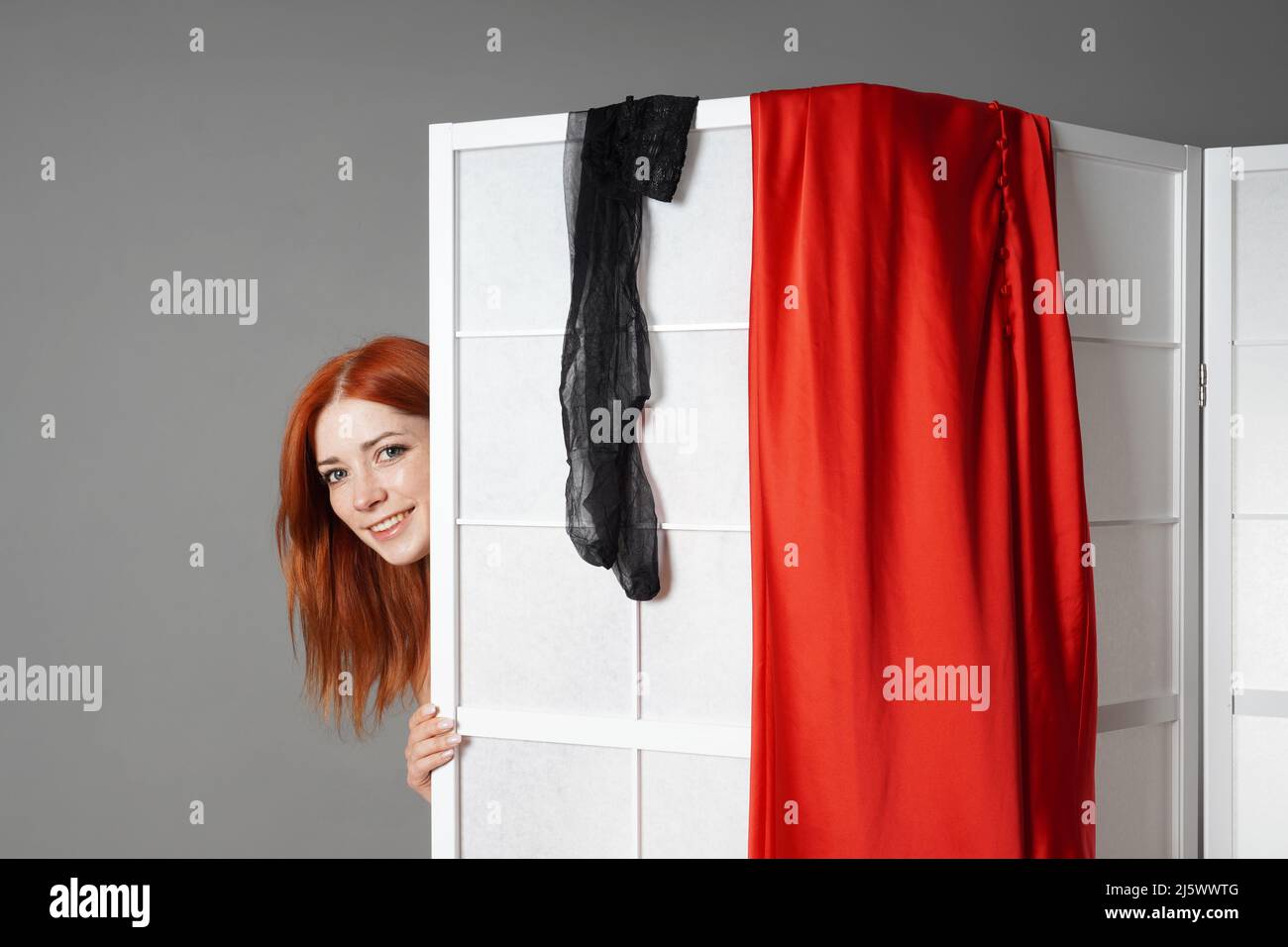 young woman peeks from behind folding screen while undressing or changing clothes Stock Photo