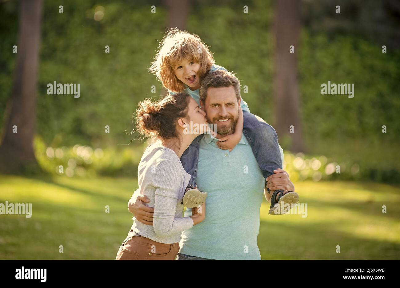Mother kiss father while boy child riding piggy back on fathers shoulders summer outdoors, family Stock Photo