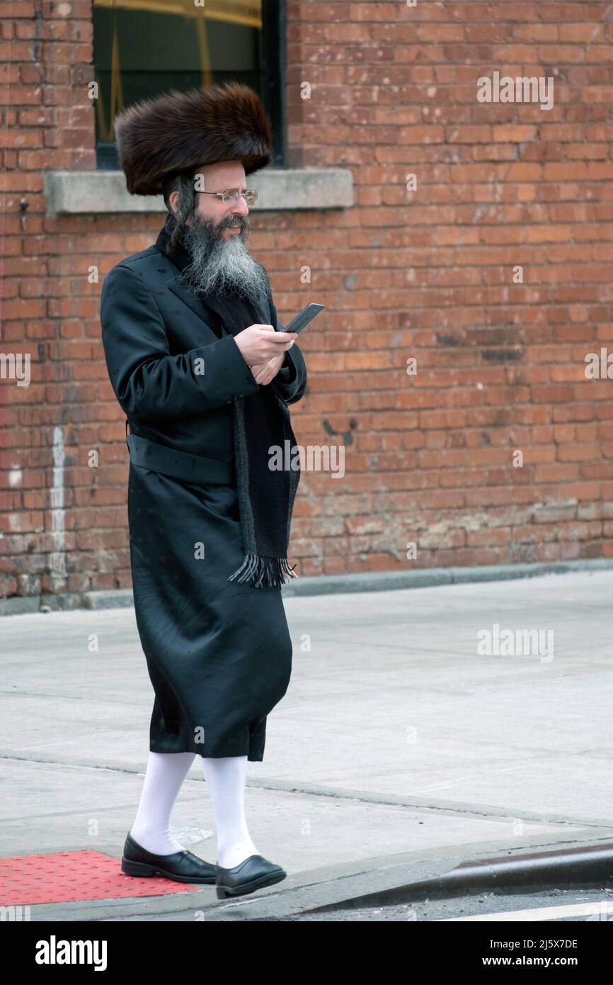 On passover and wearing long white stockings and a shtreimel fur hat, an orthodox Jewish man checks his cell phone. On Bedford Ave in Wiliamburg, NYC. Stock Photo