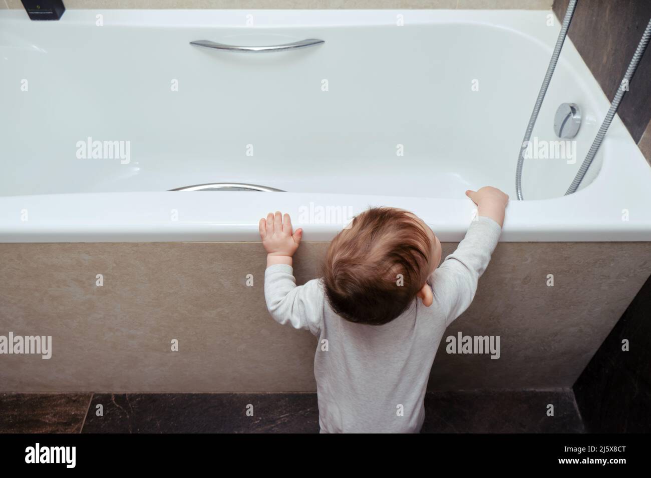 Toddler baby boy climbs into the white tub standing at the edge. Child