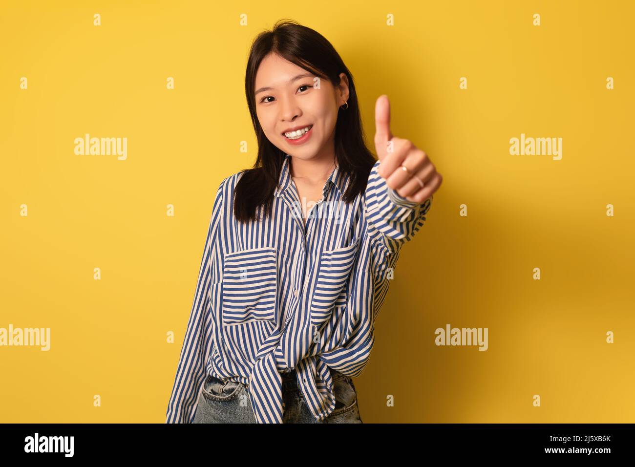 Young Asian woman showing thumb up, recommending or approving something, giving positive feedback on yellow background Stock Photo