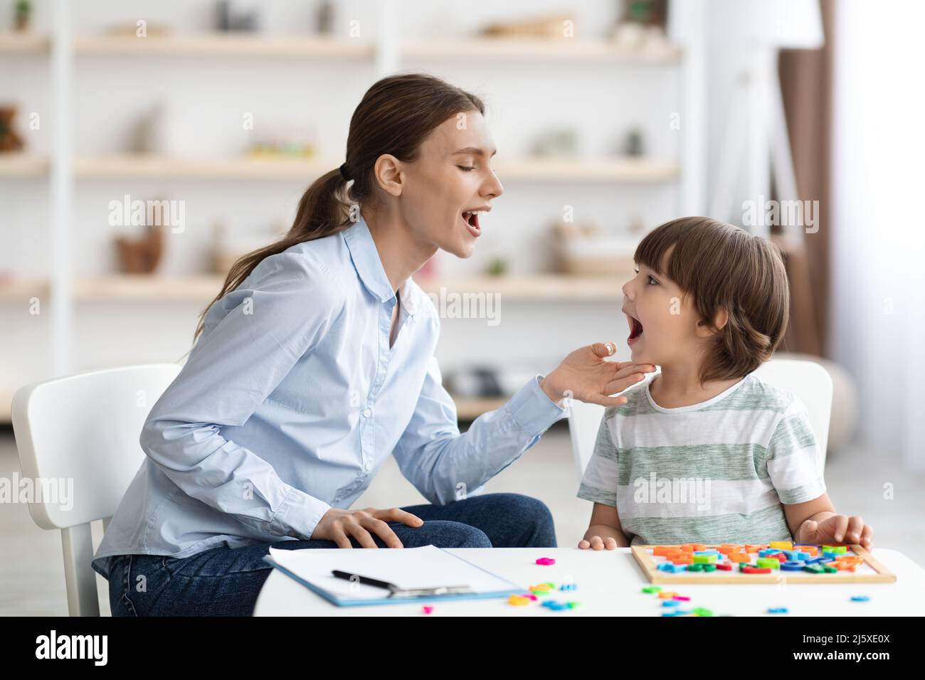 Professional woman speech therapist helping little boy to pronounce right sounds, showing mouth articulation Stock Photo