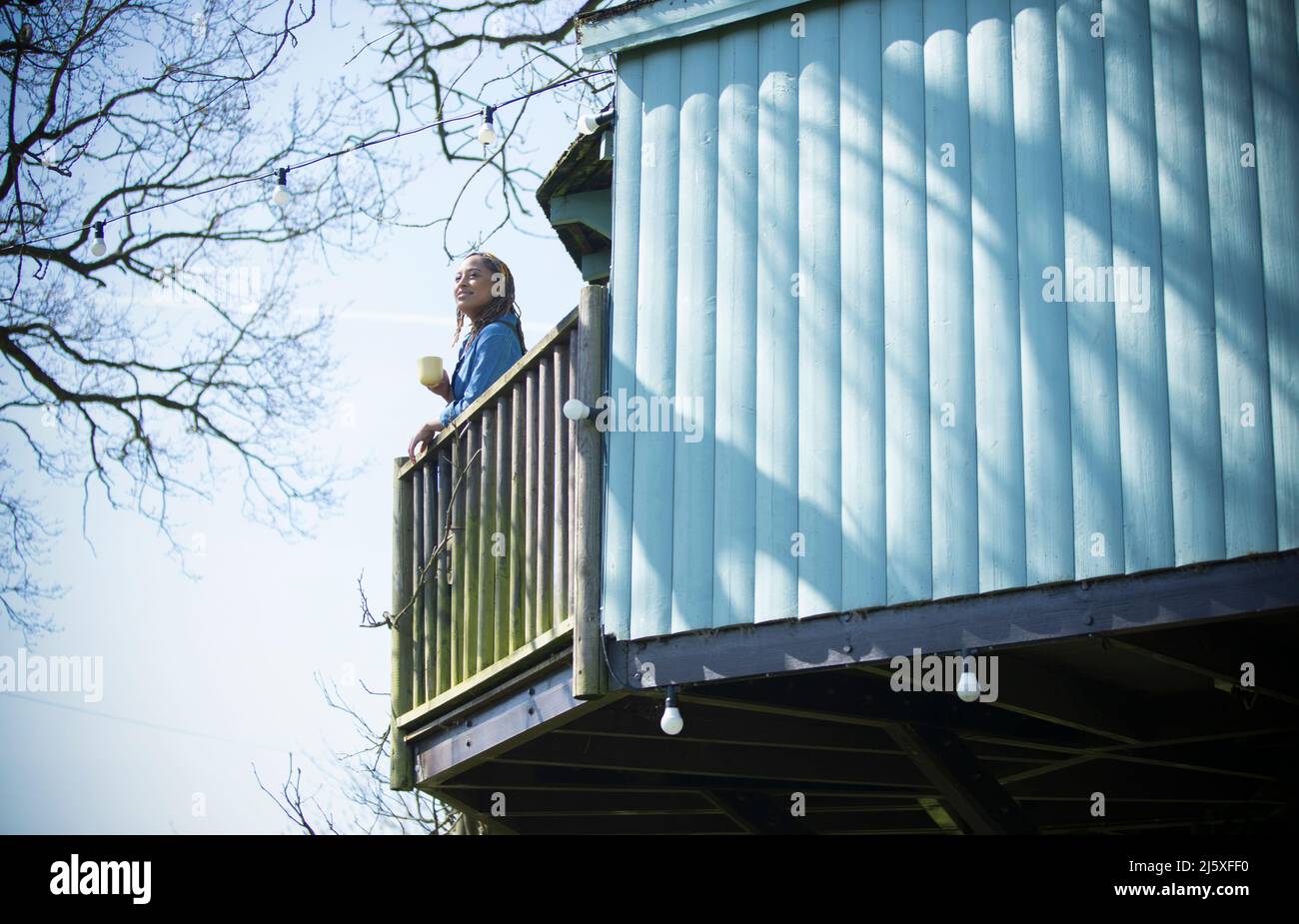 Young woman with coffee on sunny tree house balcony Stock Photo