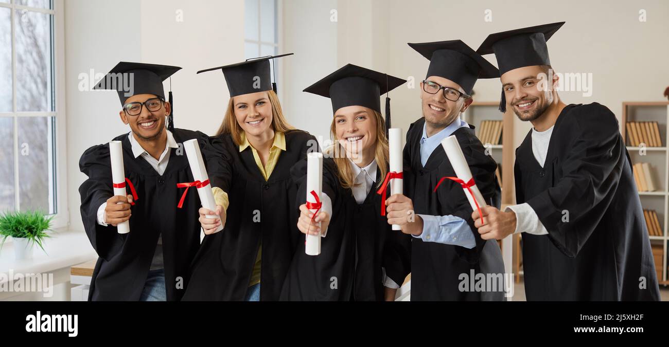 Happy college or university graduates in caps and gowns holding diploma scrolls and smiling Stock Photo