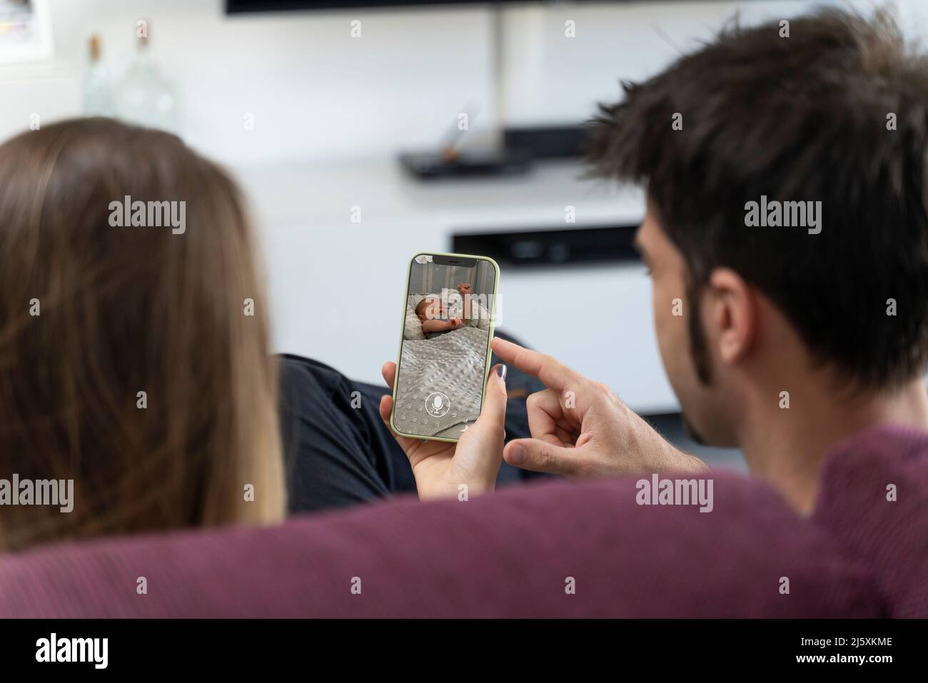 Two parents sitting on the sofa and monitoring their baby on the phone Stock Photo