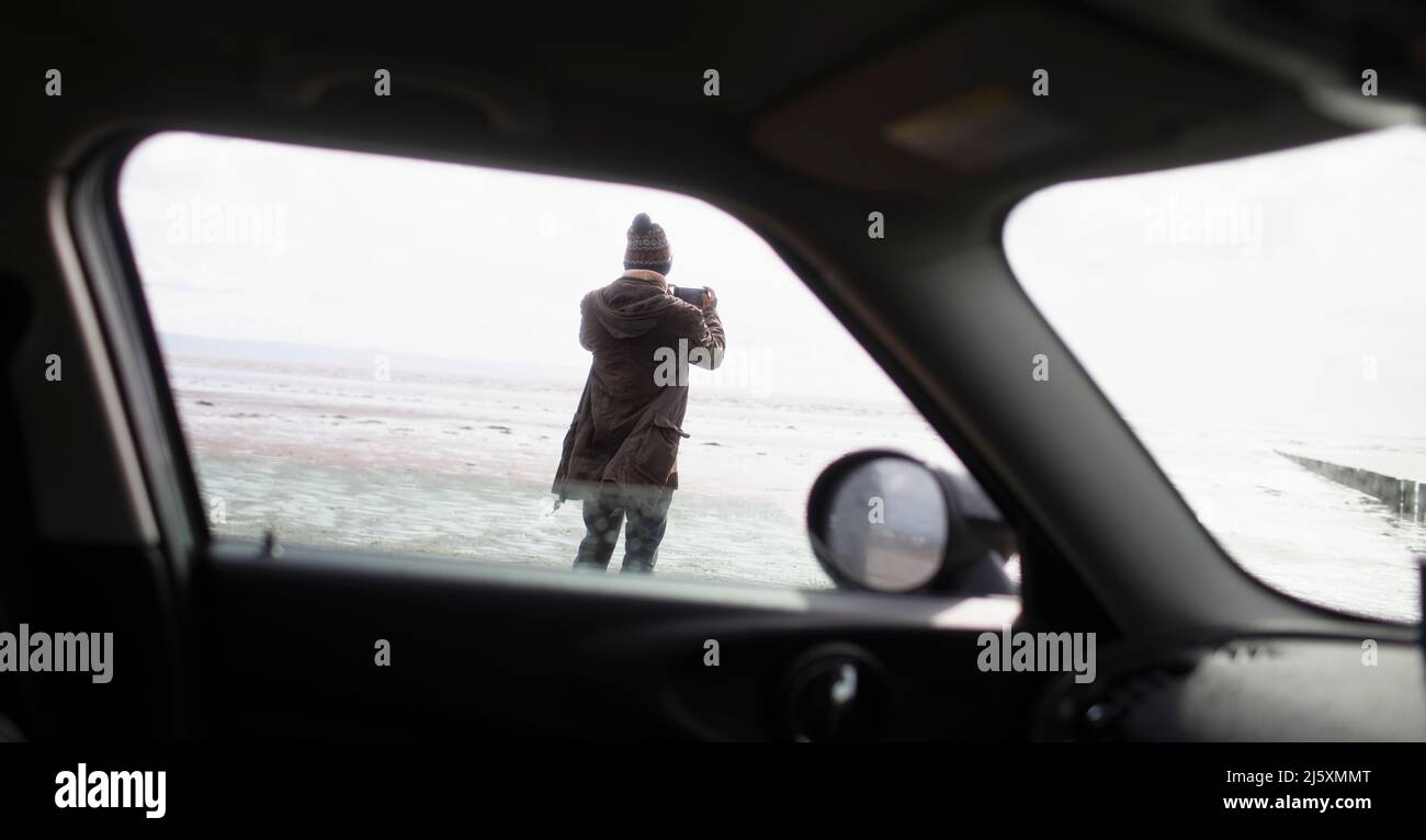 Man with camera phone on winter beach outside car Stock Photo