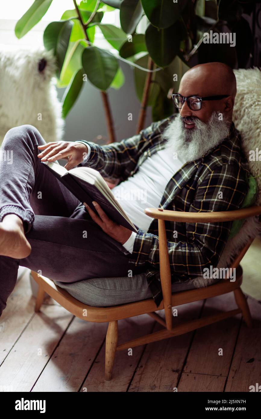 Mature man with beard reading book in armchair Stock Photo
