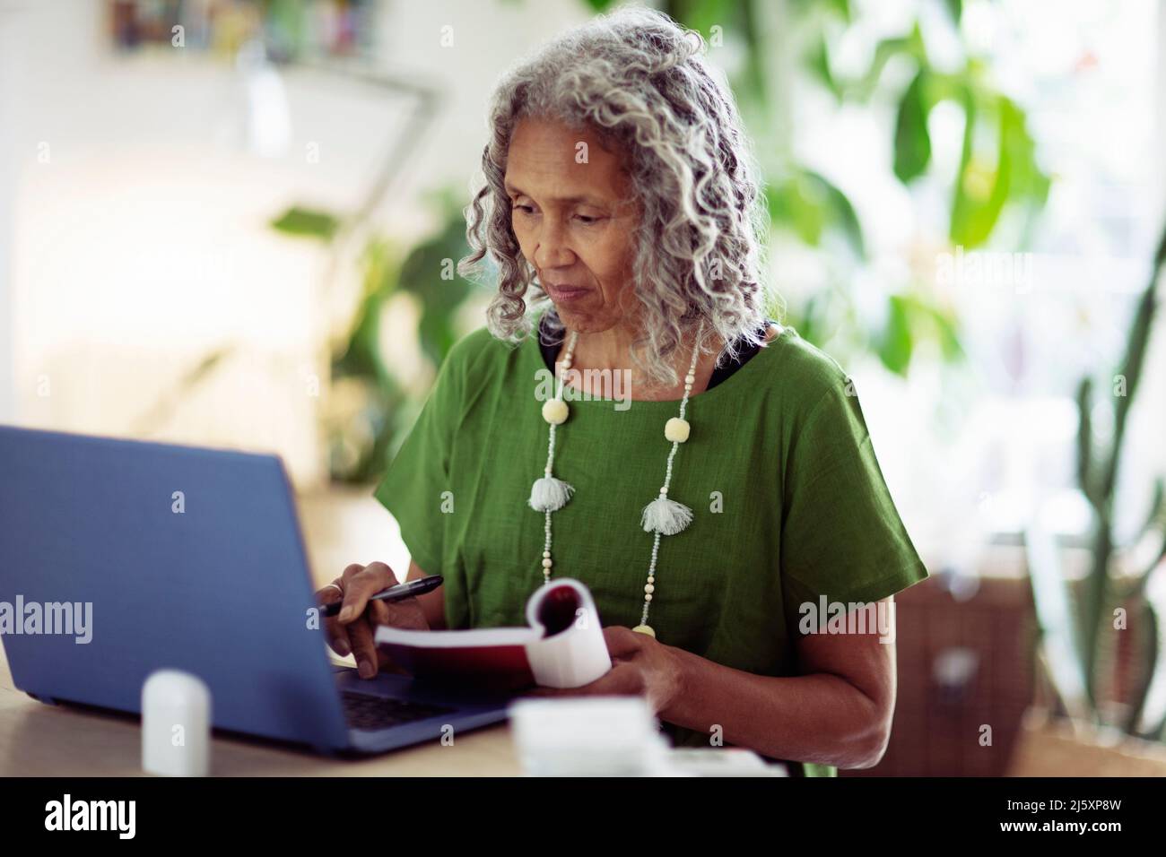 Senior woman with book working at laptop at home Stock Photo