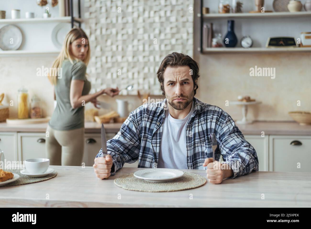 Angry hungry dissatisfied millennial caucasian man with stubble ignoring woman and wait for food in kitchen Stock Photo