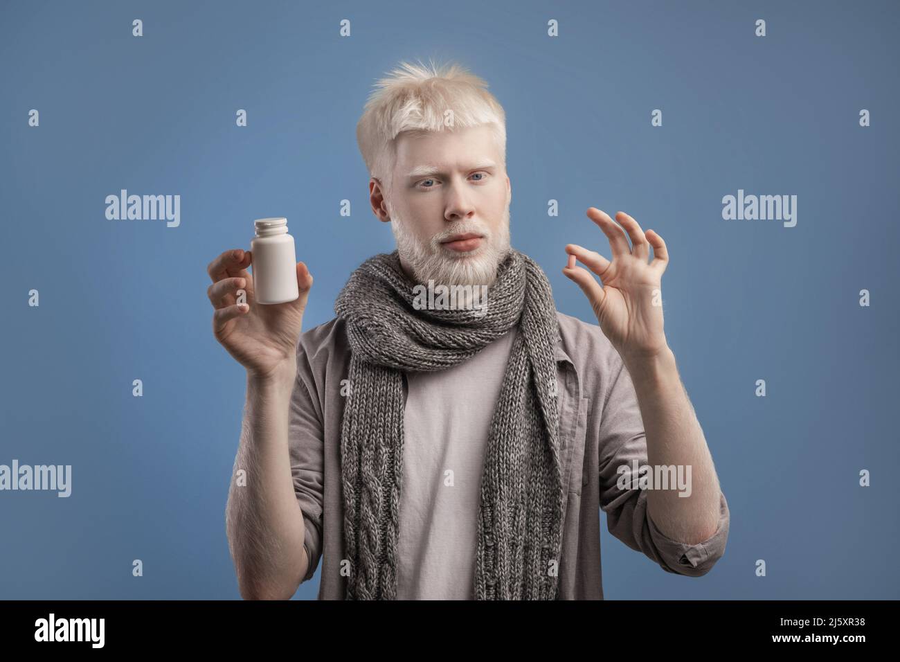 Albino guy holding jar with meds and pill in hands, standing on blue background, taking anti viral medication Stock Photo