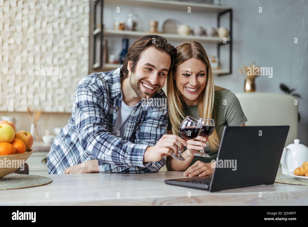 Happy young european man with stubble and lady clink glasses with wine and look at laptop in modern kitchen Stock Photo