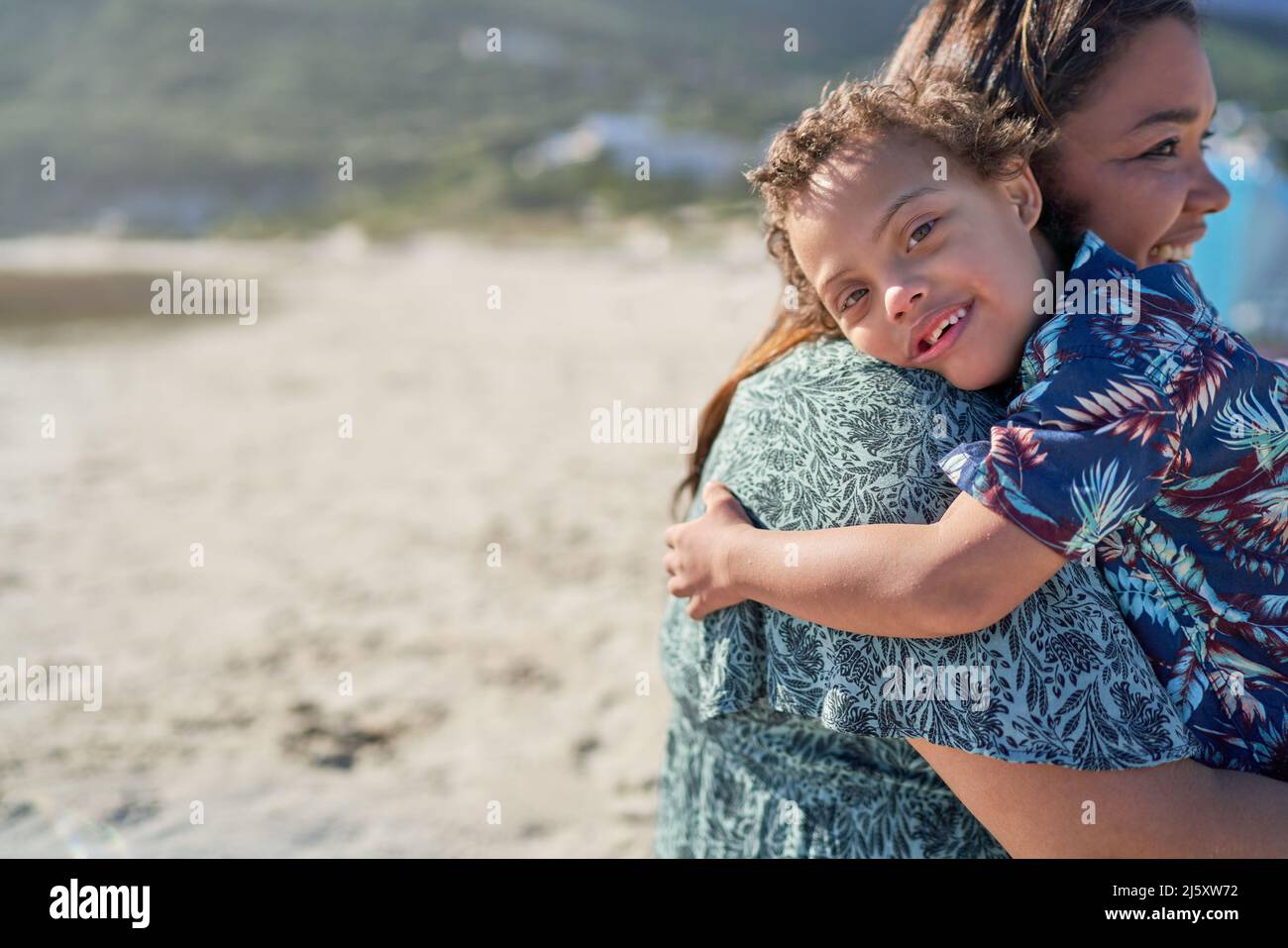 Portrait happy boy with Down Syndrome over shoulder of mother on beach Stock Photo
