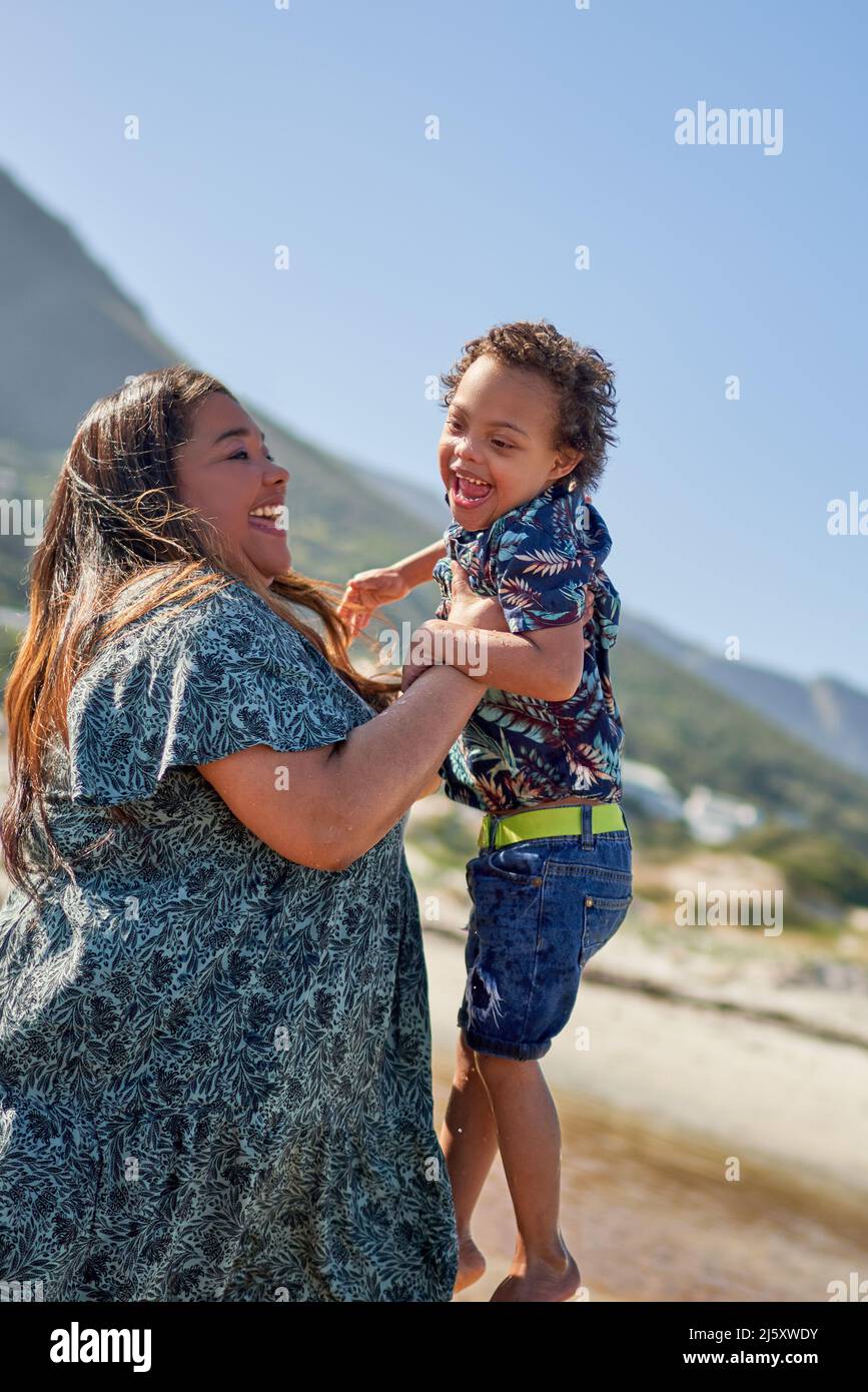 Happy mother lifting cute son with Down Syndrome on beach Stock Photo