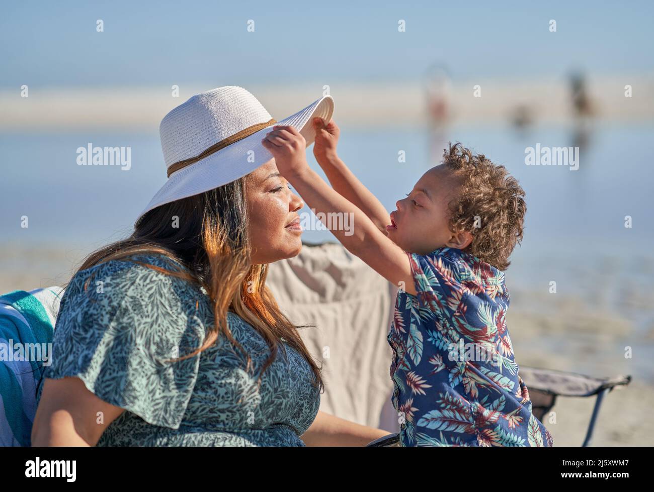 Curious son with Down Syndrome lifting mother's hat on beach Stock Photo