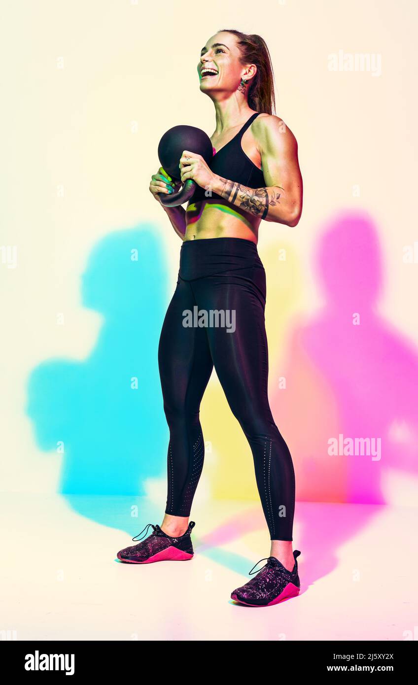 Sporty woman resting after workout with kettlebell. Photo of laughing woman in black sportswear on white background with effect of rgb colors shadows. Stock Photo