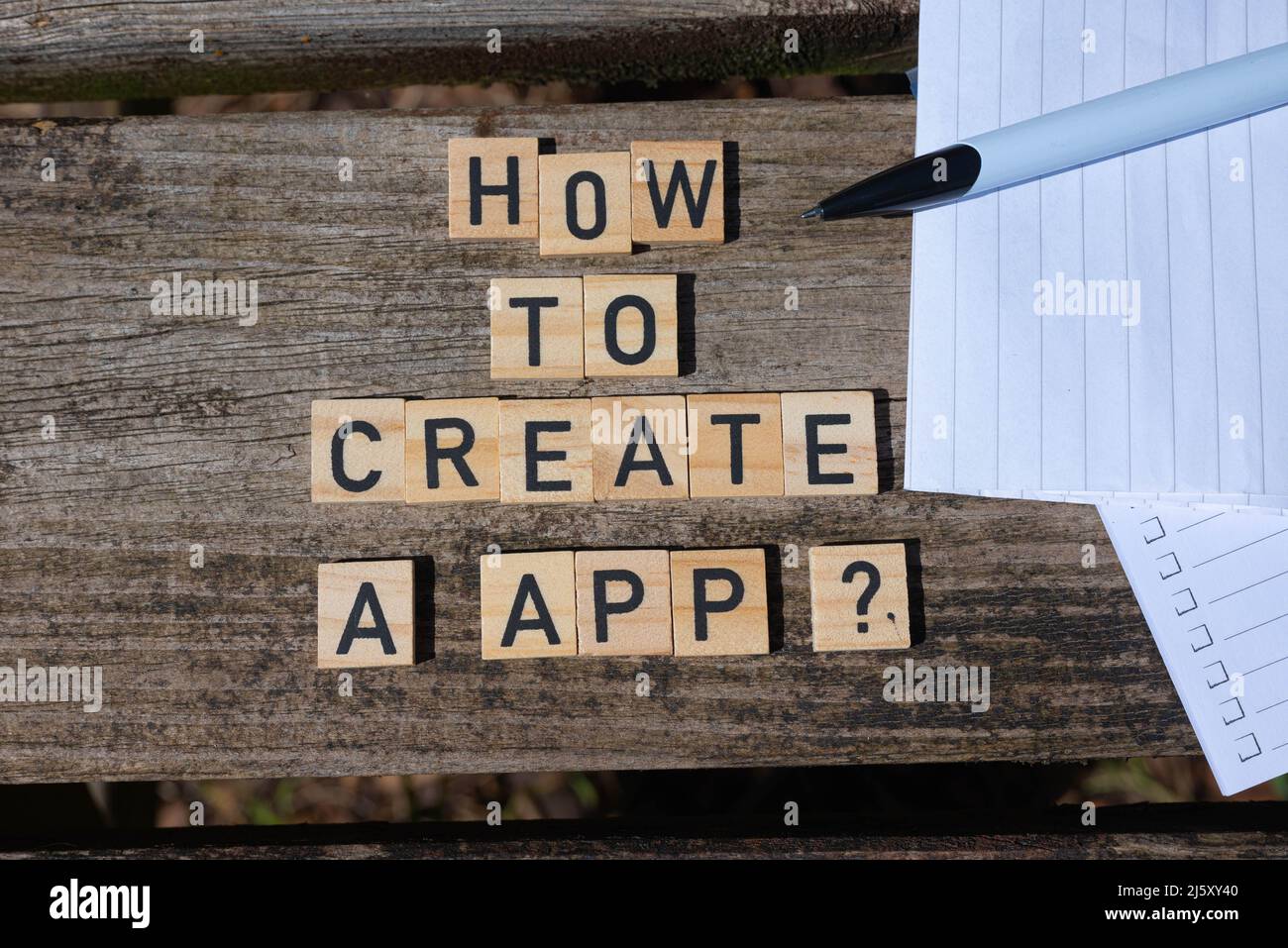 How to create a app? Black capital letter words on wooden toy blocks on a natural garden table background with paper and pencil. Perfect for websites. Stock Photo