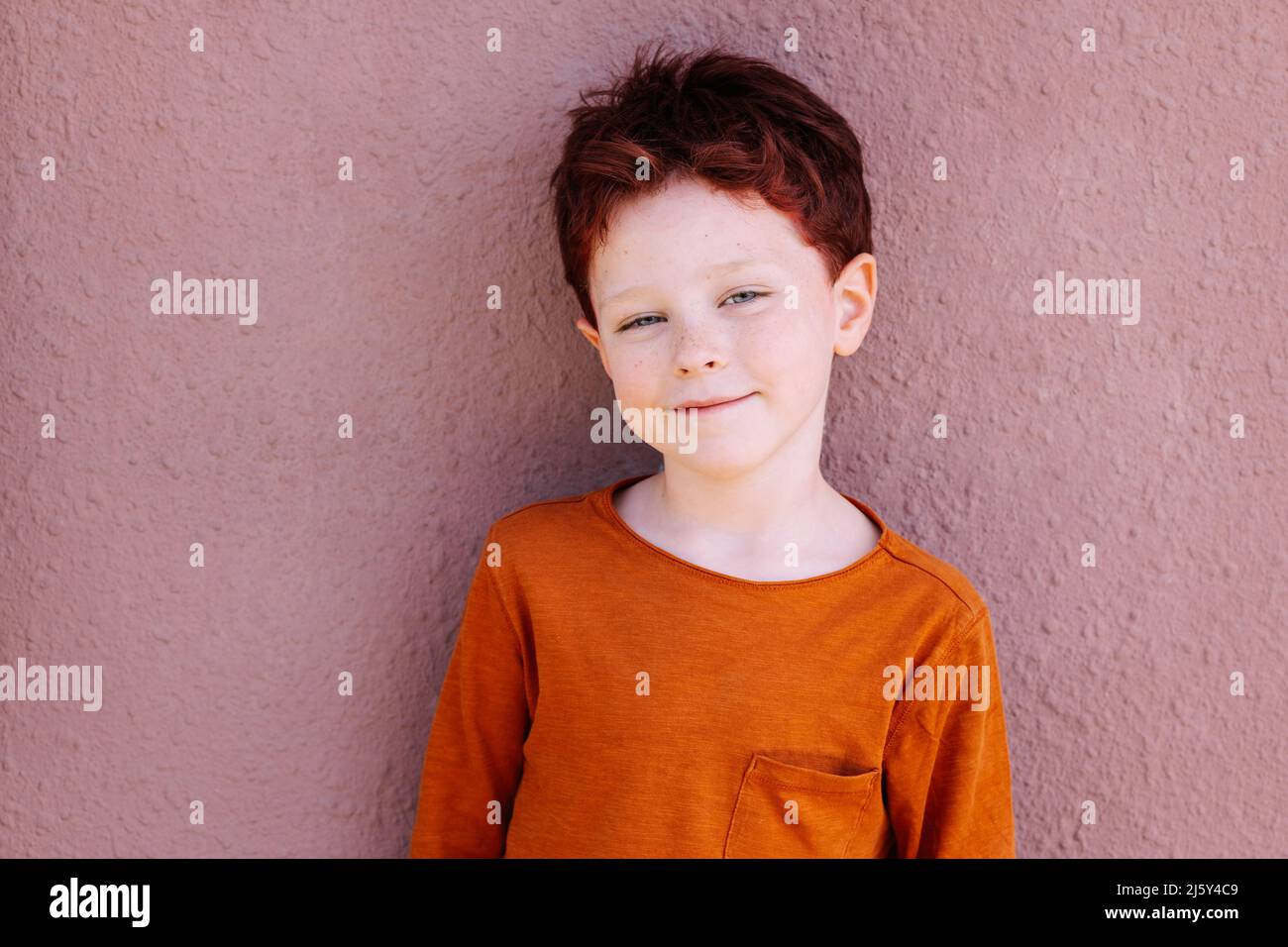 Adorable boy in casual red clothes with red hair looking at camera while standing against pink background on street of city Stock Photo
