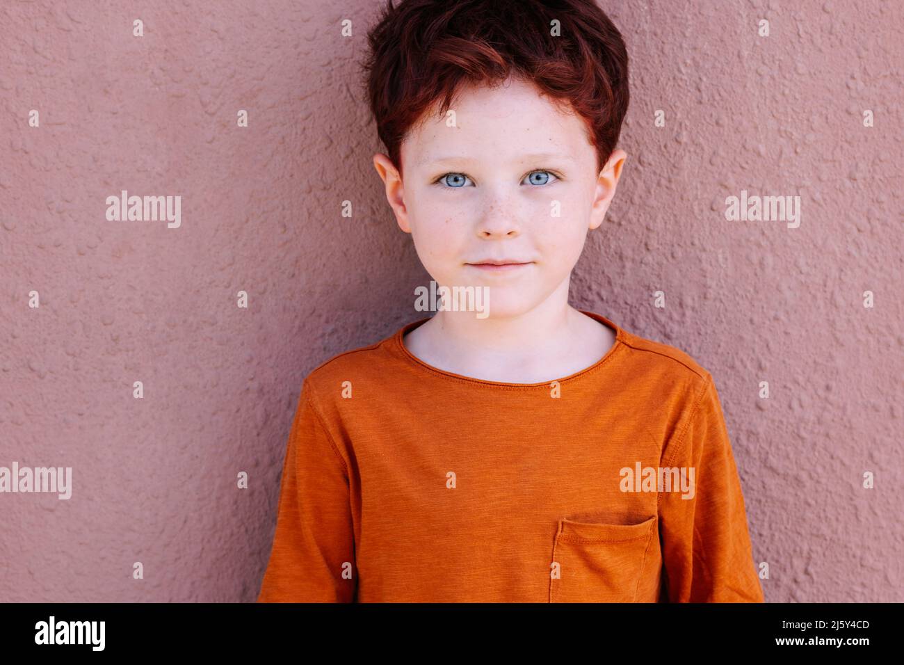 Adorable boy in casual red clothes with red hair looking at camera while standing against pink background on street of city Stock Photo
