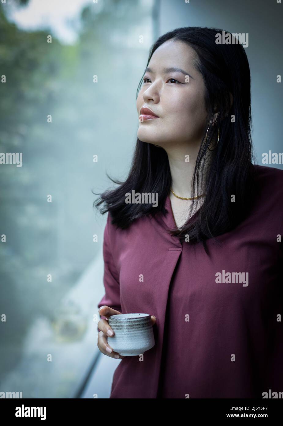 Thoughtful young woman drinking tea at window Stock Photo