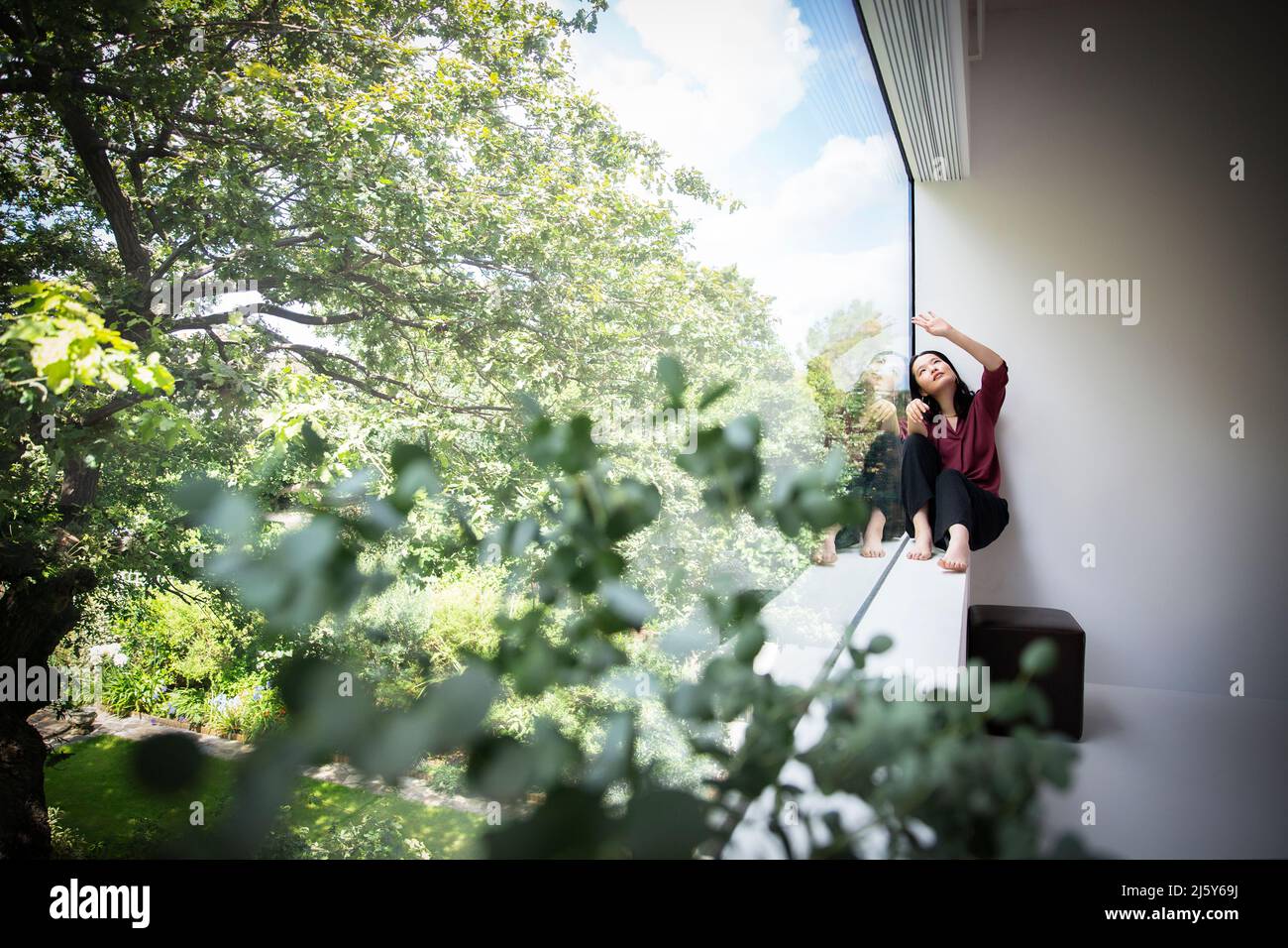 Young woman sitting in window with view of green trees Stock Photo
