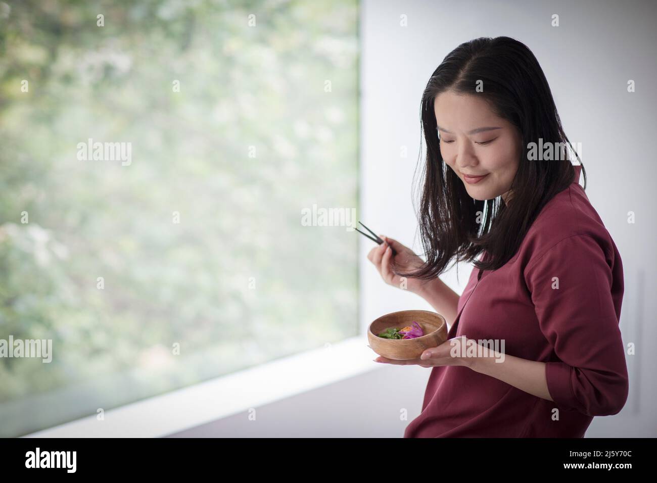 Young woman eating from bowl with chopsticks at window Stock Photo