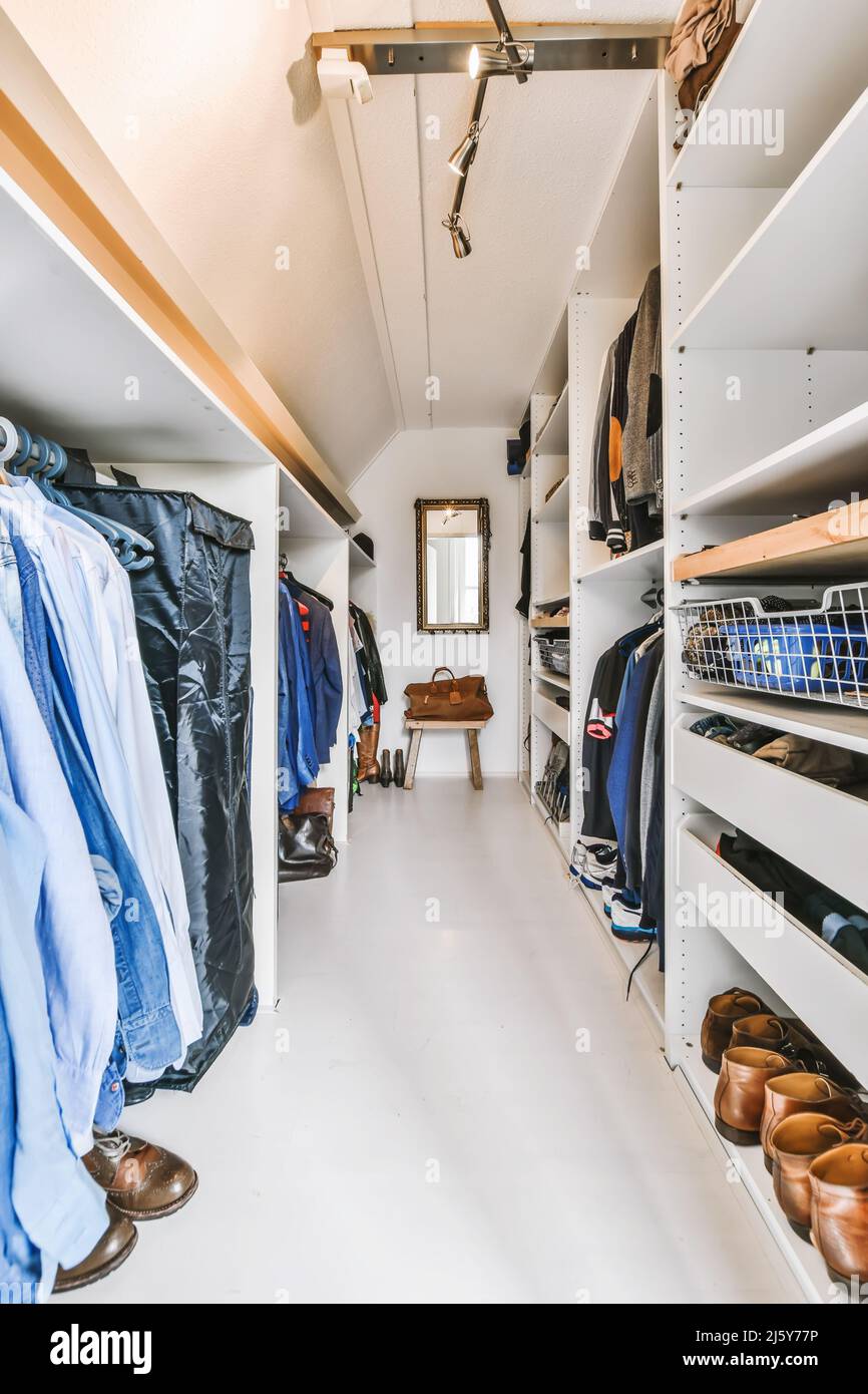 Long light wardrobe with abundance of stylish garments hanging on racks and arranged on white shelves against mirror in apartment Stock Photo