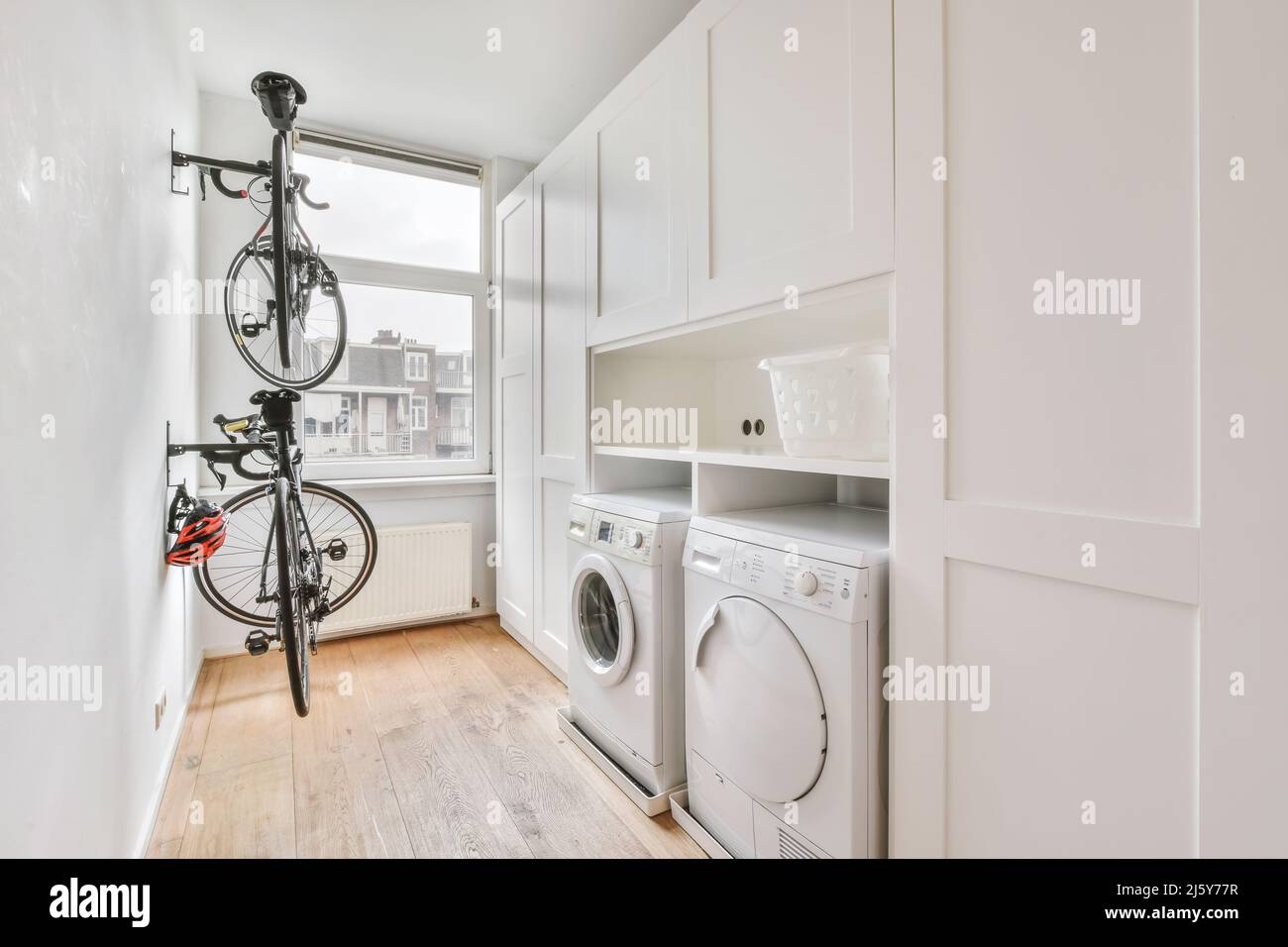Modern white electric washing and dryer machines placed near white cabinets and basket in light laundry room with window and bicycle Stock Photo