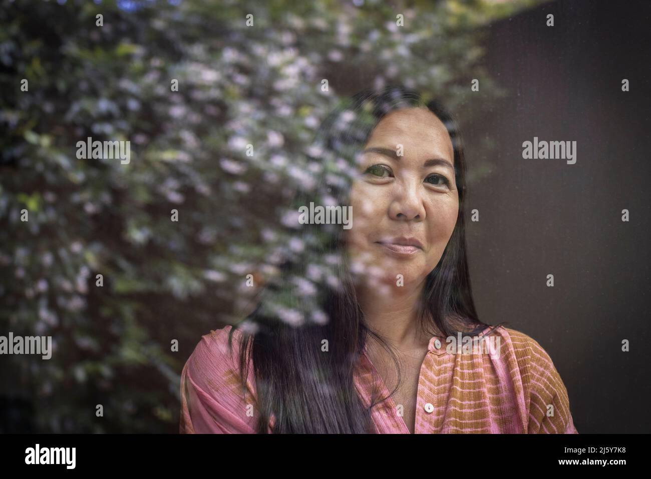 Portrait confident woman at window with reflection of tree Stock Photo
