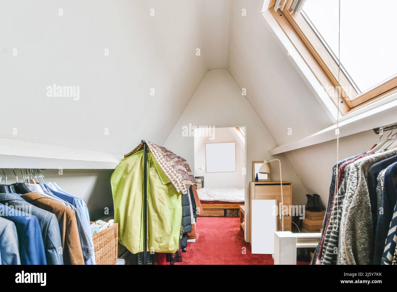 Collection of various elegant suits and outerwear hanging on racks near window in light attic with wooden drawer in apartment Stock Photo
