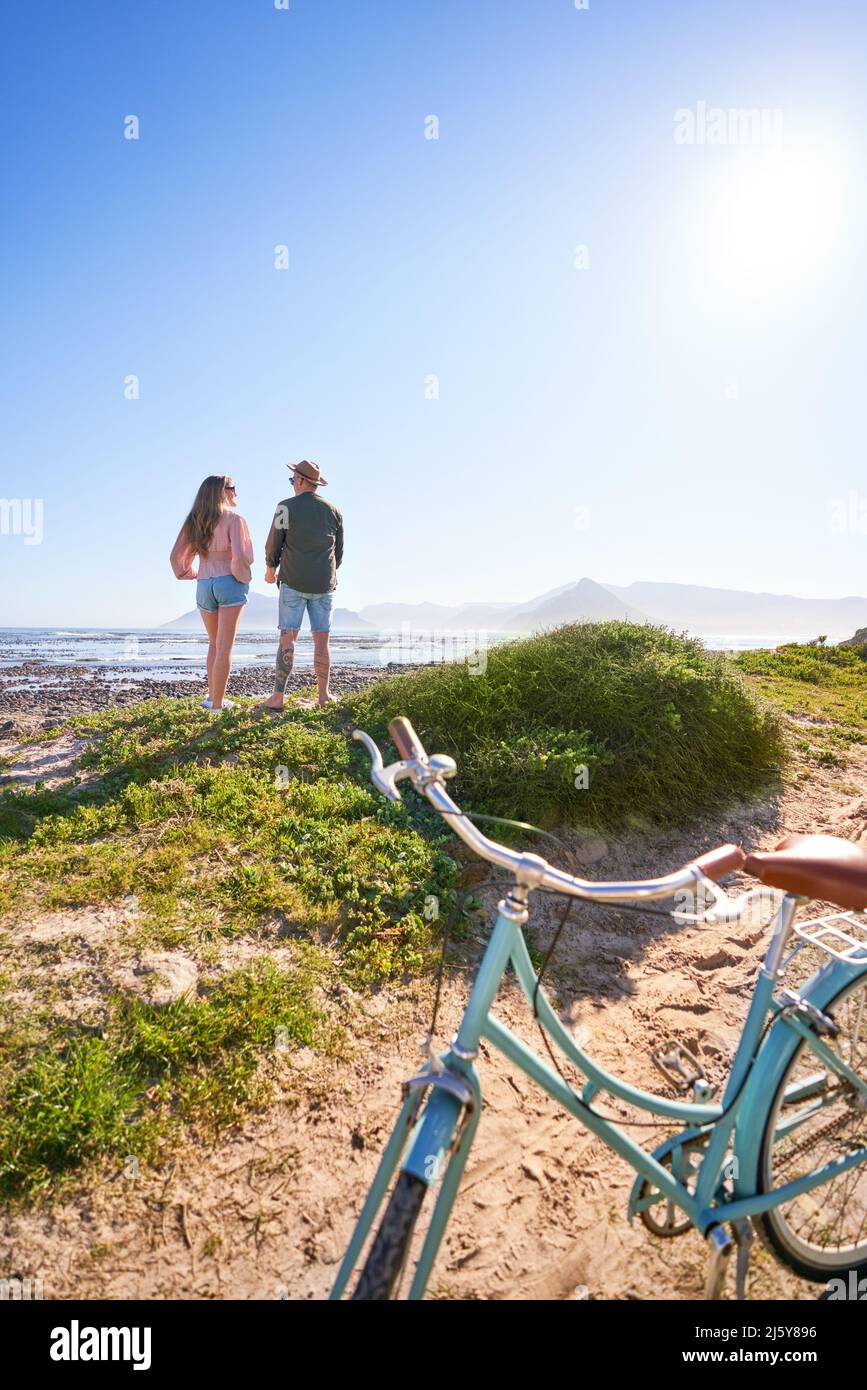 Couple enjoying sunny summer ocean view from beach behind bicycle Stock Photo
