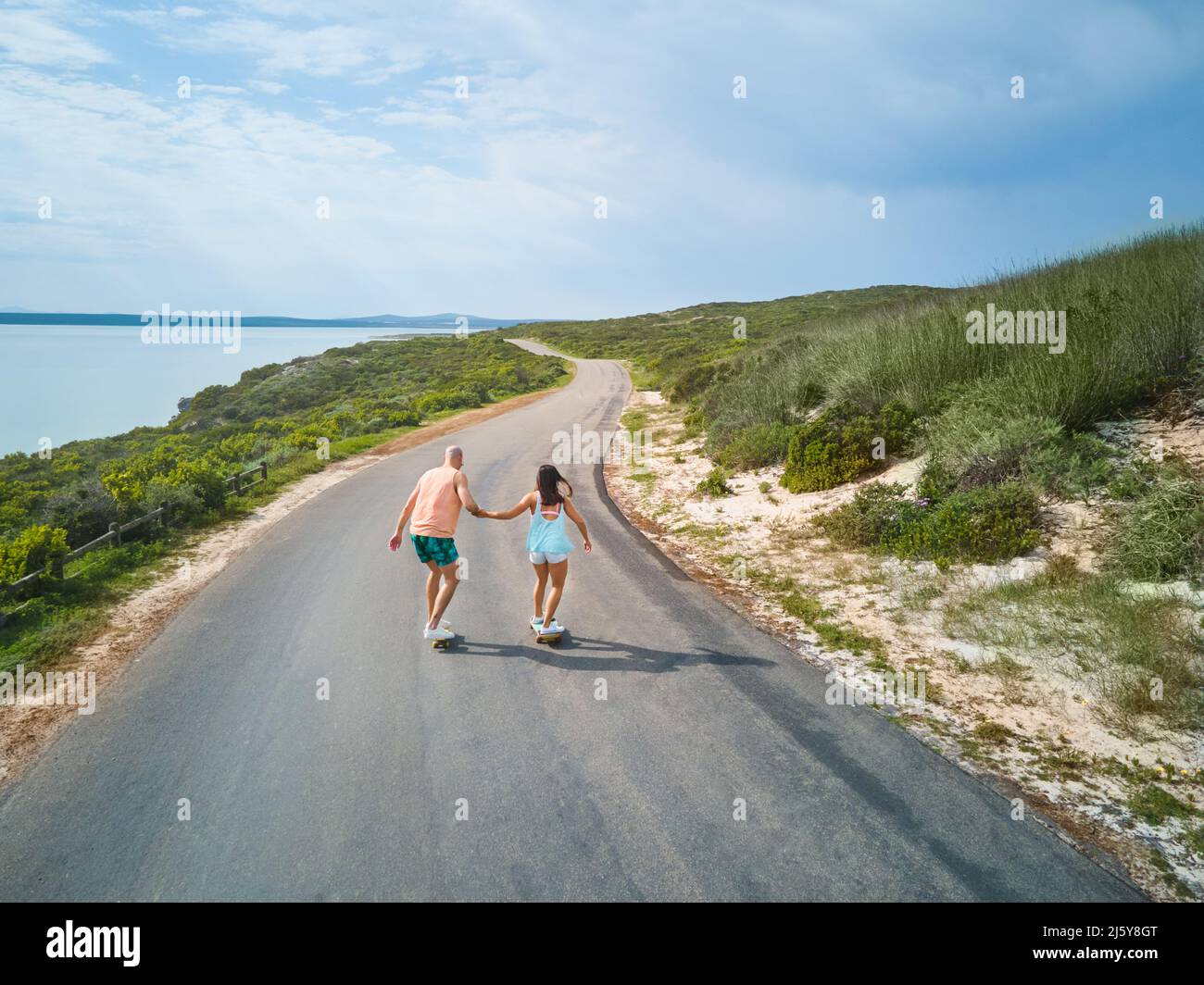 Couple holding hands and skateboarding on sunny ocean road Stock Photo