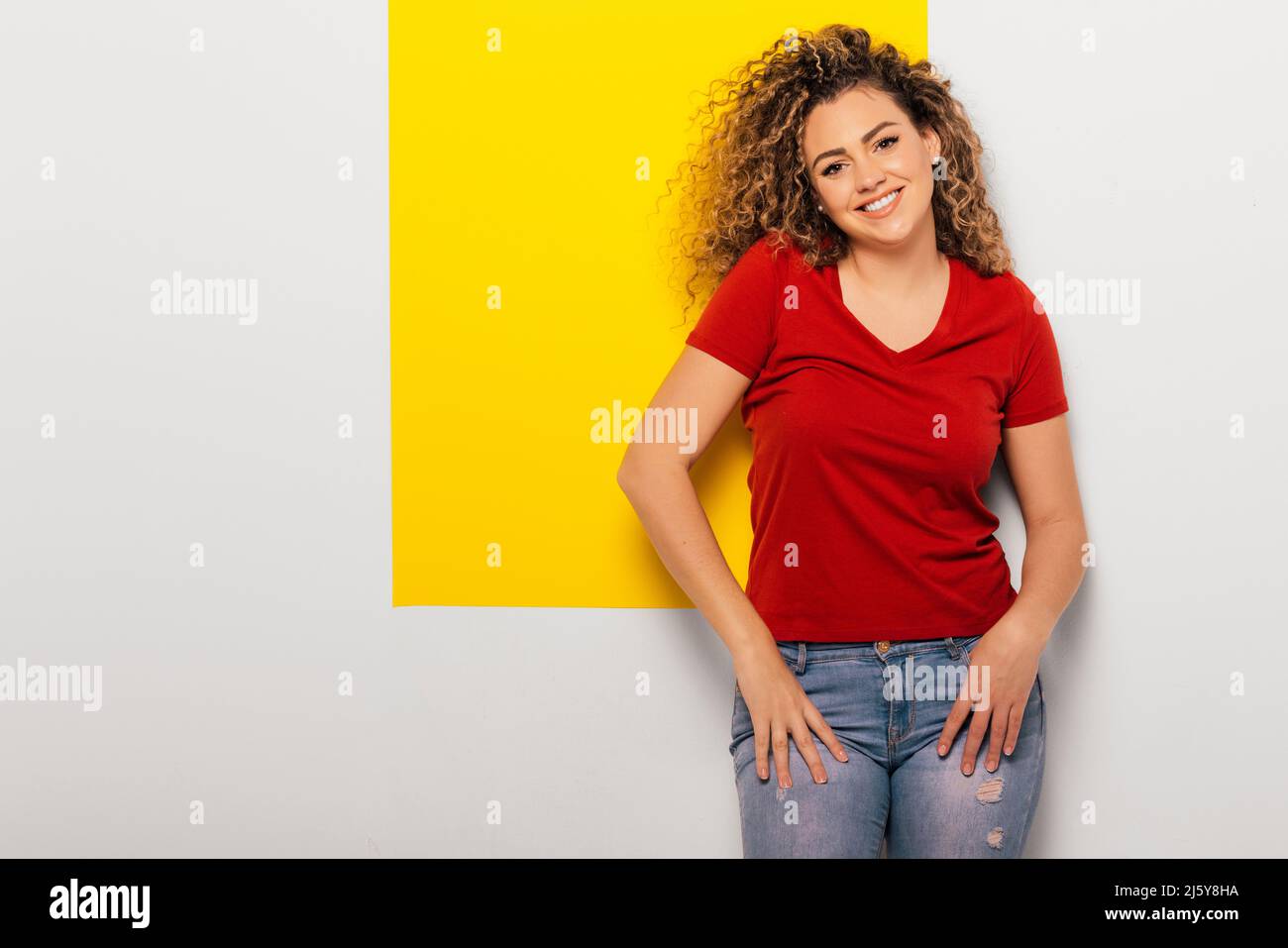 Cheerful young Woman in red t-shirt and jeans looking at camera on white and yellow background Stock Photo