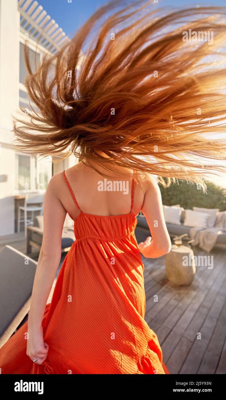 Carefree woman in orange dress flipping long red hair on sunny patio Stock Photo