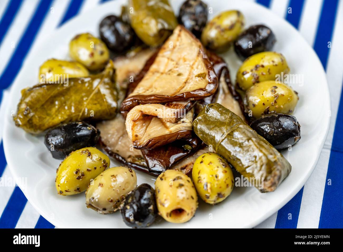 Greek appetizer plate with olive, sliced eggplant and dolmades (vine leaf stuffed with rice) on plate on blue striped towel, closeup. Stock Photo