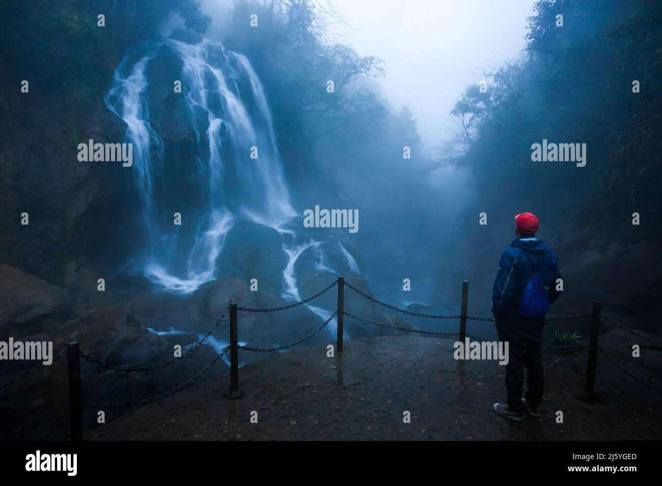 A tourist man wearing a red hat stands in front of a tropical waterfall in foggy, a tourist attraction in Sapa, Vietnam. Long exposure. Stock Photo