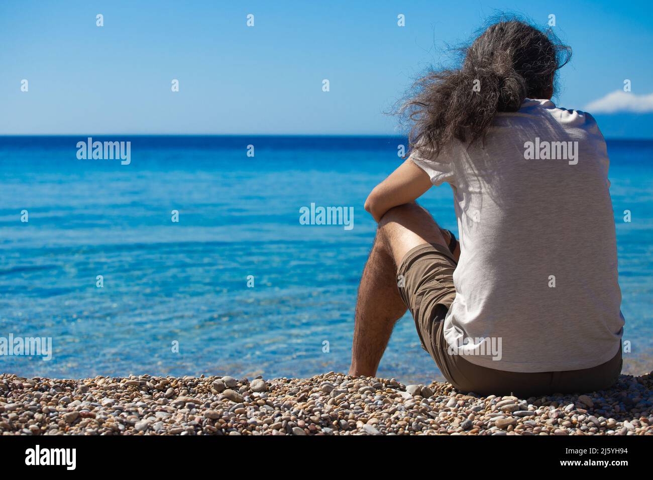 Man is sitting lonely from the back and looking to the Mediterranean blue sea. Horizontal. Stock Photo