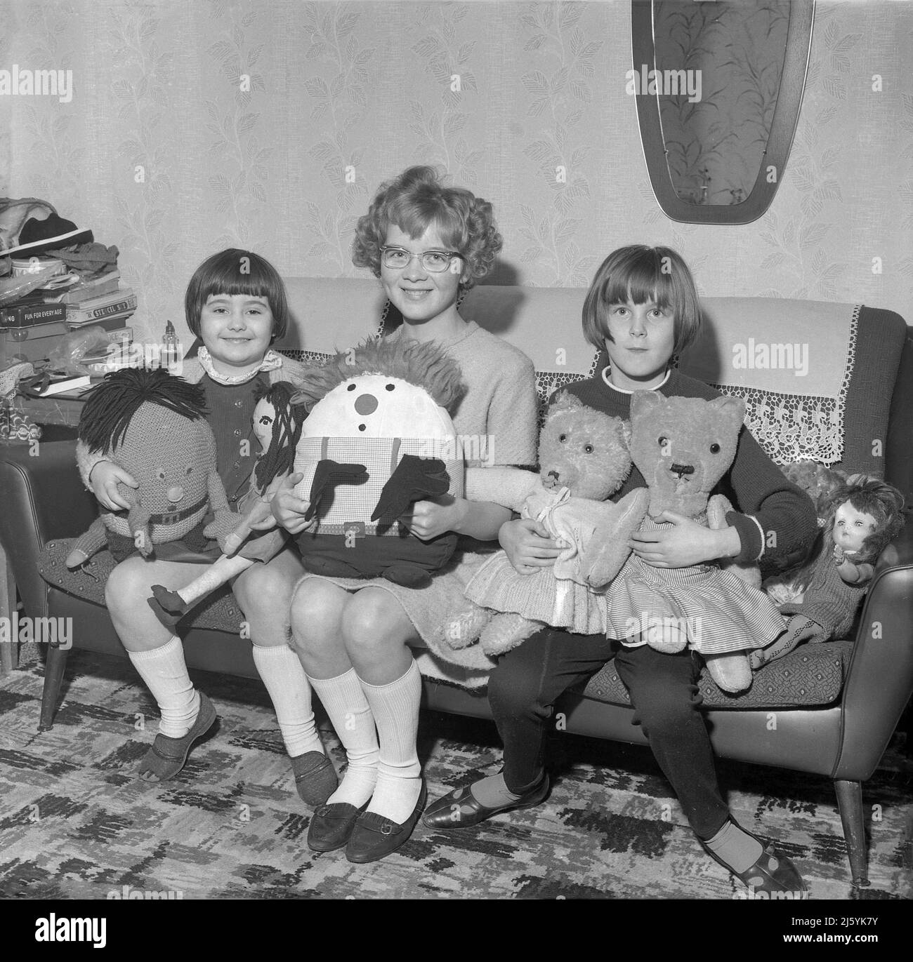 1966, historical, three young girls sitting on a sofa of the era, holding their precious soft toys, ragdolls and teddy bears, England, UK. Stock Photo