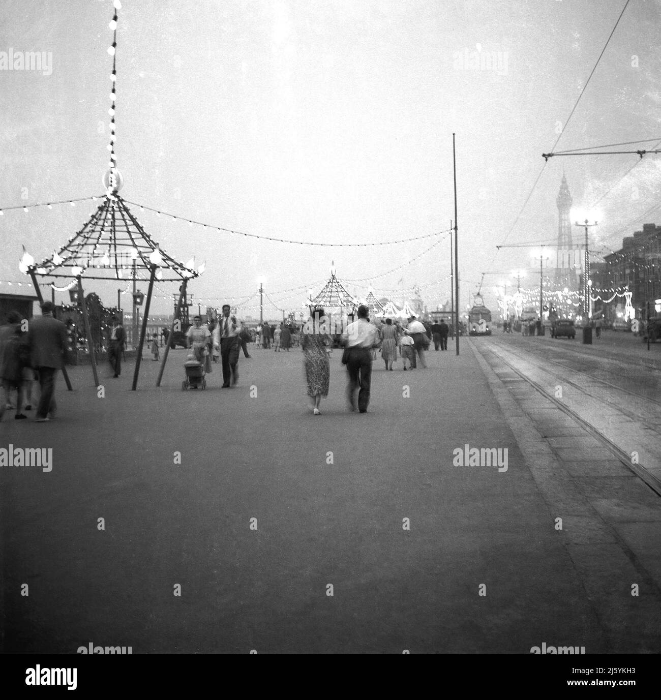 1960, historical, early evening and people stroll along the front at Blackpool, England, UK. The tram line is on the right of the promenade, while the town's famous tower can be seen in the distance. Stock Photo