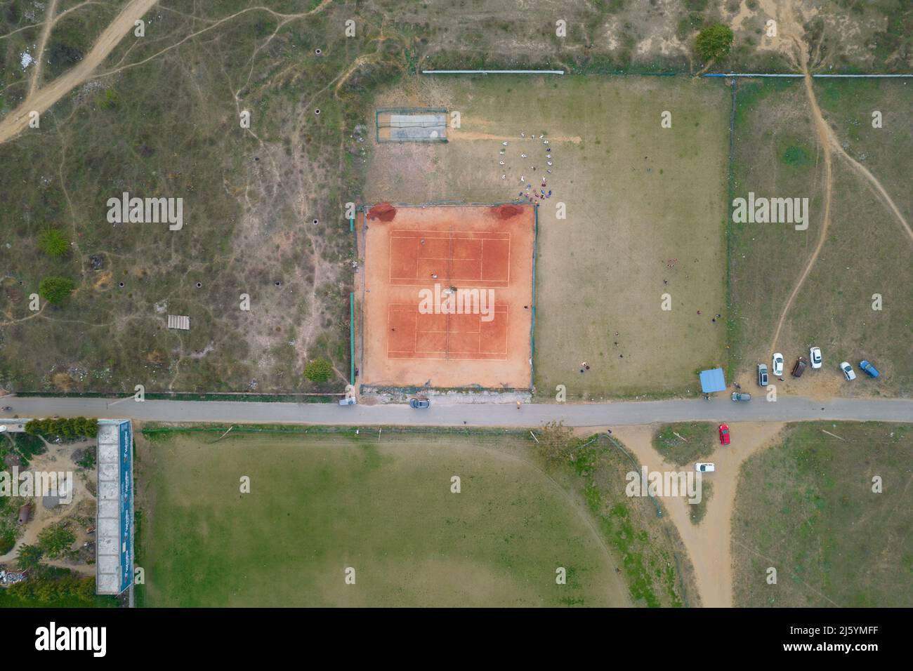 top down aerial drone shot showing clay tennis court and cricket pitch with children people practicing sitting in a circle in rural urban area in Stock Photo