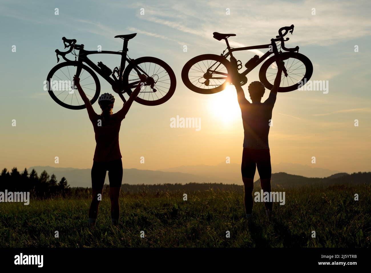 Two cyclers silhouettes holding road bicycles high above heads, in winner pose during sunset over the mountain landscape Stock Photo