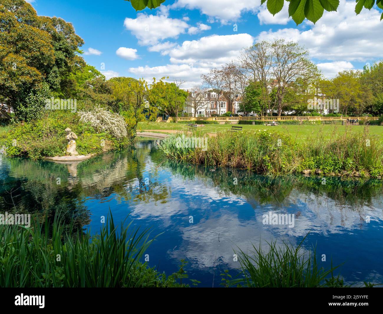 A broad landscape view of the sky reflected in the pond and fresh green vegetation in Fulham park in London Stock Photo