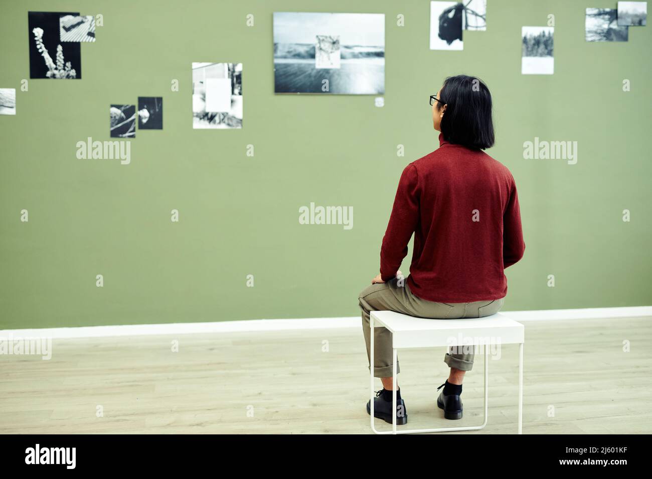 Rear view of unrecognizable young man sitting on stool in modern art gallery looking at black and white photos on green wall Stock Photo