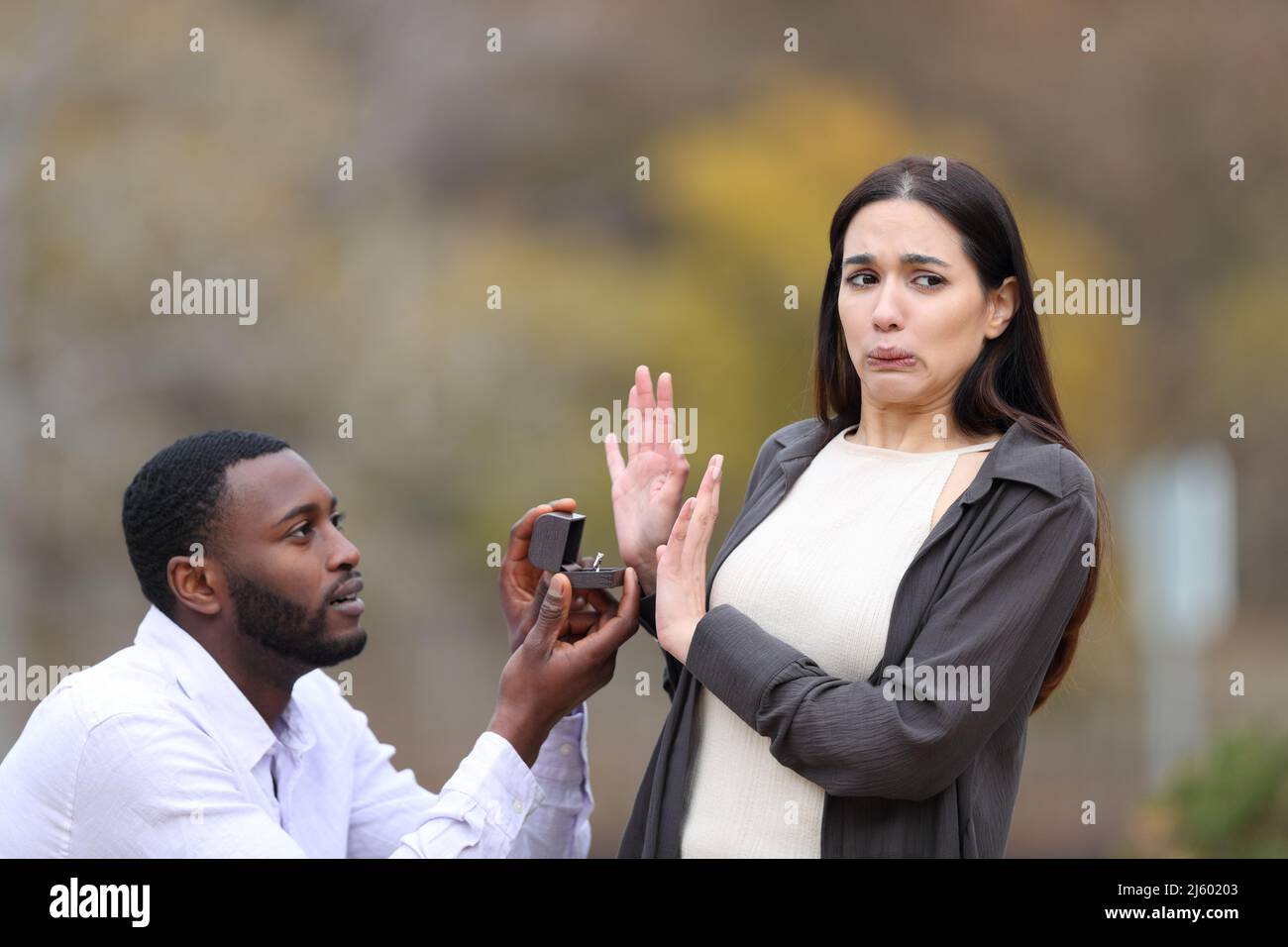 Stressed man with black skin proposing marriage and scared caucasian woman rejecting him in a park Stock Photo