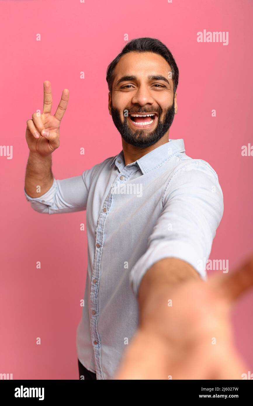 Portrait of middled-aged Indian handsome man taking selfie, looking at camera and showing V-gesture with hand, point of view of photo. Indoor studio shot isolated on pink background, vertical Stock Photo