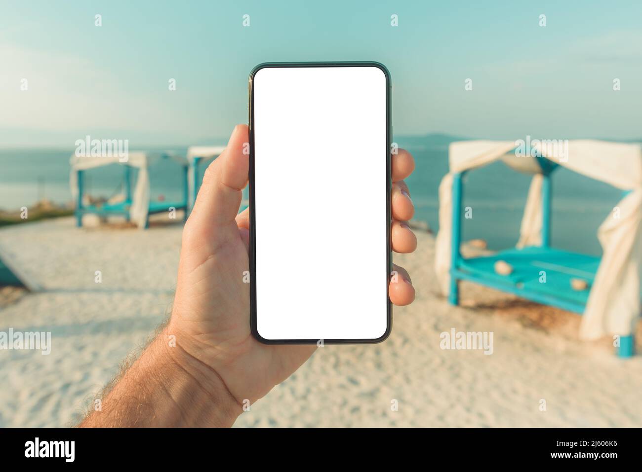 Mobile smart phone with blank mockup template screen in male hand, beach pergola in background, selective focus Stock Photo