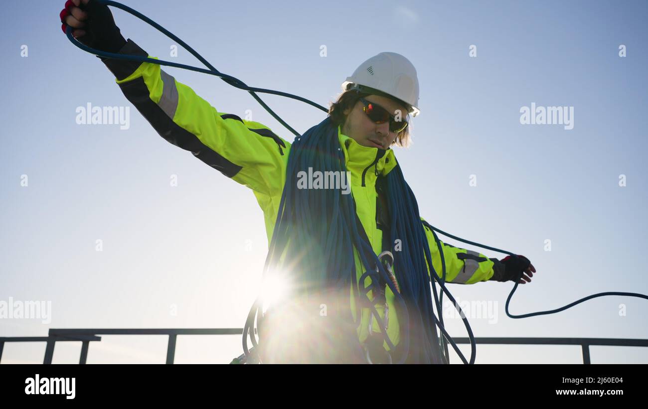 Industrial climber in a white helmet, glasses and light green overalls coils a rope on the roof against the backdrop of the sun Stock Photo