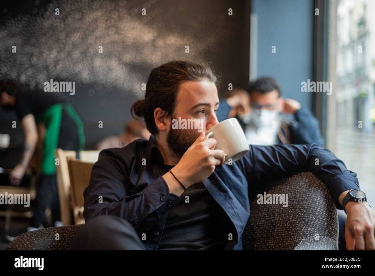Long hair Hispanic young adult male drinking in a mug at hipster coffee shop Stock Photo