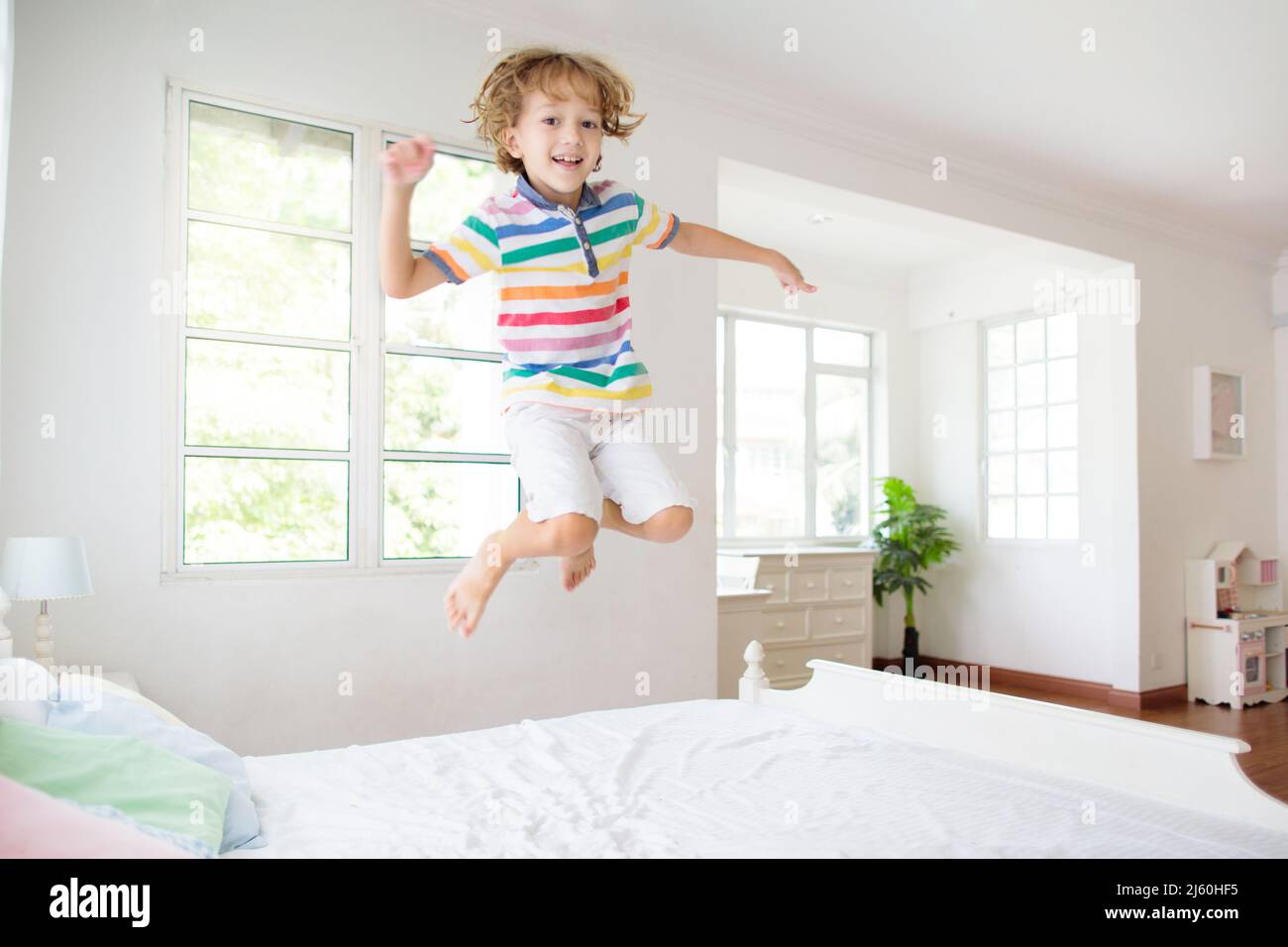 Child jumping on bed in white bedroom with big window. Kids play at