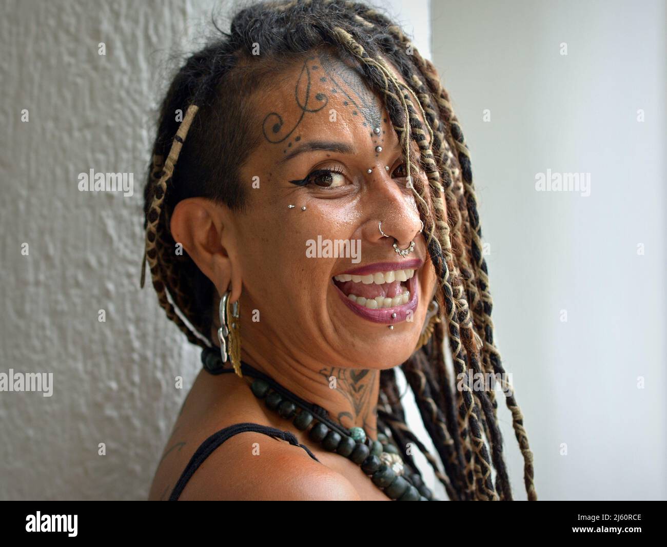 Attractive alternative young Mexican woman with long dread Rasta braids, body art facial piercing and tattoo laughs positively optimistic at viewer. Stock Photo
